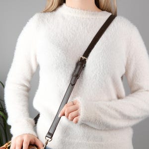 以下が含まれることがあります： ゴールドカラーのバックルと金具が付いた、濃い茶色の革製ハンドバッグストラップ。 ストラップは模様入りのハンドバッグに取り付けられています。 画像の下のテキストは次のとおりです: バッグは販売されていません。