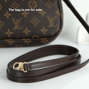 May include: A dark brown leather purse strap with gold-colored hardware. The strap is coiled on a white surface. The purse in the background has a brown monogram pattern and the text "The bag is not for sale".