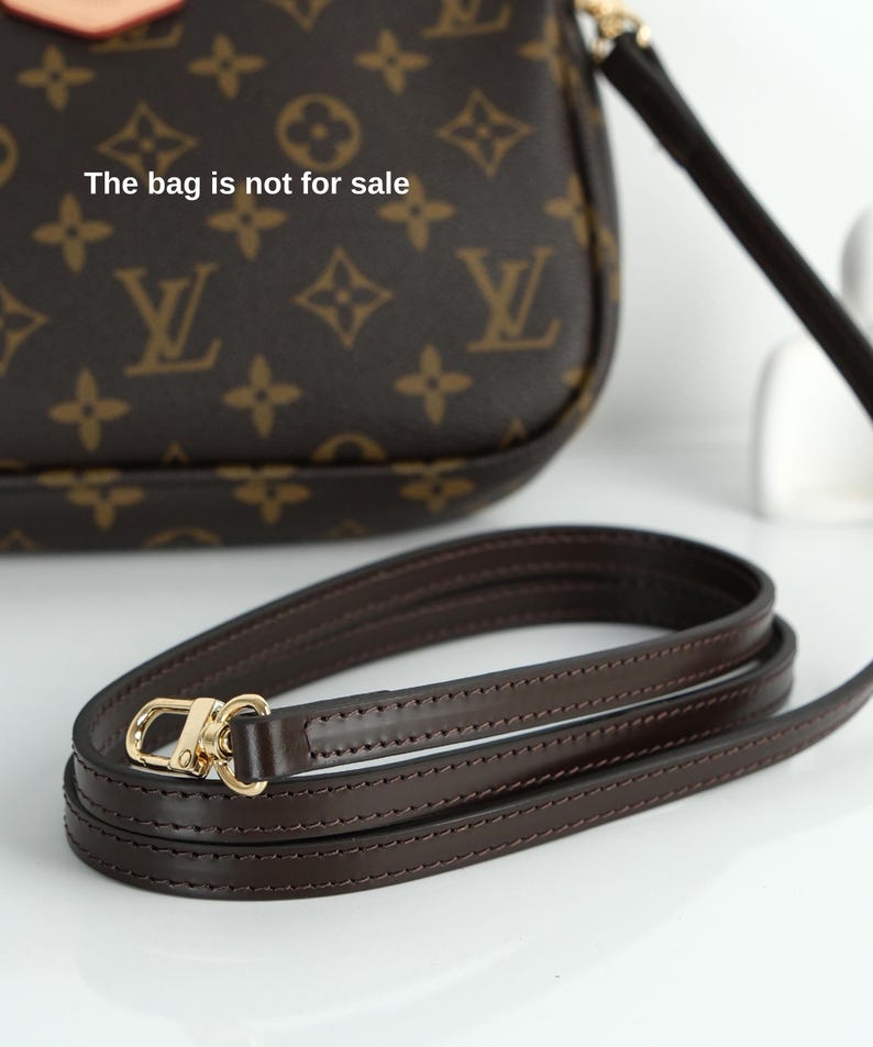May include: A dark brown leather purse strap with gold-colored hardware. The strap is coiled on a white surface. The purse in the background has a brown monogram pattern and the text "The bag is not for sale".