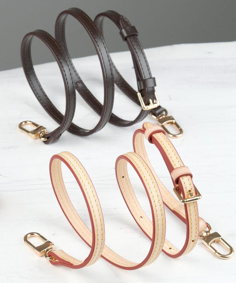 May include: Two leather purse straps. One is dark brown with gold-coloured hardware. The other is light beige with red edges and gold-coloured hardware. Both straps have a buckle and a clasp.