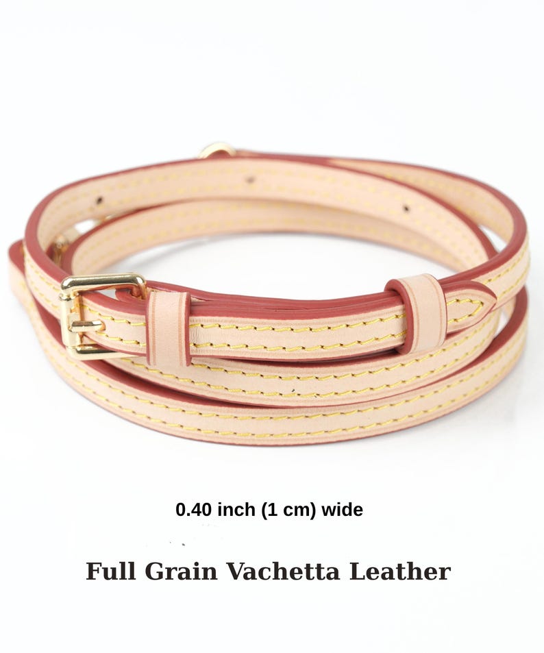 May include: A coiled leather strap in a light hue, with a gold-coloured buckle. The strap is 1 cm wide and has red edges and gold stitching. The text at the bottom reads "Full Grain Vachetta Leather".