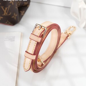 May include: A beige leather strap with a gold buckle and a red leather lining. The strap is folded and coiled, and the buckle is visible in the center of the image. The bag is not for sale.