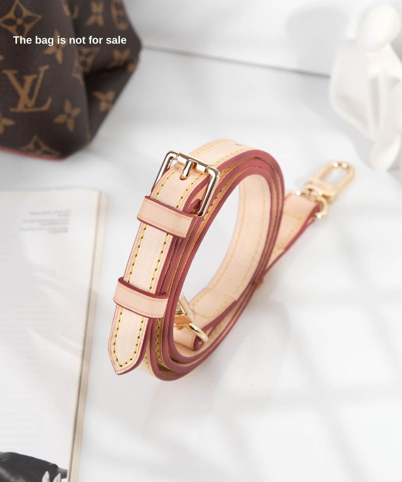 May include: A beige leather strap with a gold buckle and a red leather lining. The strap is folded and coiled, and the buckle is visible in the center of the image. The bag is not for sale.