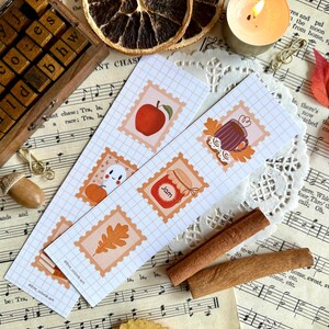 Könnte beinhalten: Zwei dekorative Lesezeichen mit herbstlichen Illustrationen, darunter ein Apfel, eine Tasse und ein Glas Marmelade. Die Lesezeichen liegen auf einem Hintergrund aus Notenblättern, mit Zimtstangen und einer brennenden Kerze.