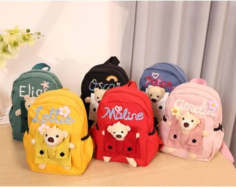 May include: Six colorful mini backpacks with embroidered teddy bears and names. The backpacks are green, black, blue, yellow, red, and pink.