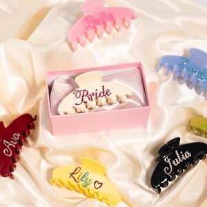 May include: A collection of personalized hair claw clips in various colors, including pink, red, yellow, black, and blue. Each clip features a name or word, such as "Bella," "Ava," "Lily," "Julia," and "Bride," embellished with rhinestones. The clips are displayed on a satin surface.