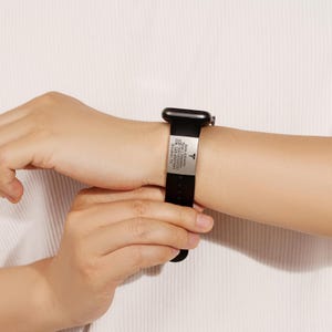 May include: Black Apple Watch band with a silver medical ID tag engraved with medical information, including allergies, medications, and emergency contact information. The tag also features a medical symbol.