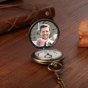 May include: Vintage bronze pocket watch with a chain. The watch face has black numerals on a white background. The open lid shows a photograph of a smiling person. The watch is on a wooden surface.