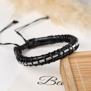 May include: Black leather bracelet with silver-colored metal accents. The bracelet features a woven design and adjustable ties. The jewelry is displayed on a wooden tray.