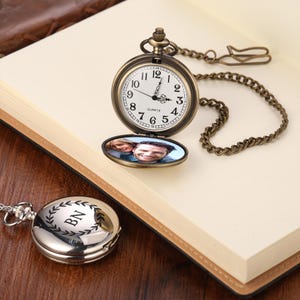 Può includere: Un orologio da tasca in stile vintage con finitura bronzo, aperto per rivelare una foto. Il quadrante mostra numeri romani e lancette che indicano l'una. È visibile anche un orologio da tasca argentato con le iniziali "BN".
