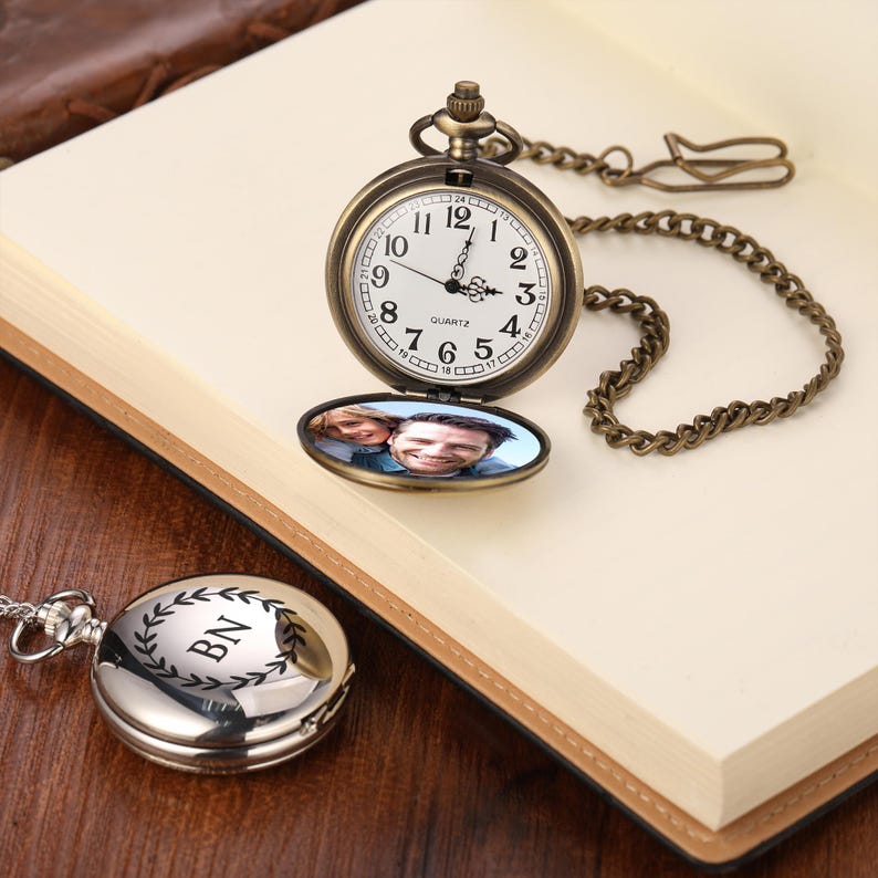 May include: A bronze-coloured pocket watch with a chain, open to show a photograph. The watch face has Roman numerals and hands at 1 o'clock. A silver pocket watch with the initials "BN" is also visible.