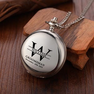 May include: A silver pocket watch with a chain, engraved with the name "WILLIAM" and the word "GROOMSMAN" along with a date. The watch is round and has a polished finish. The watch is resting on a wooden surface.