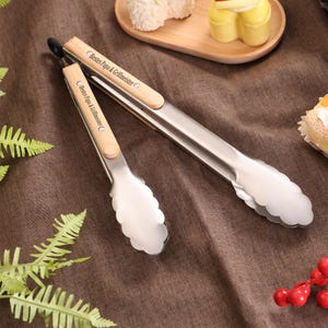 May include: Two stainless steel tongs with wooden handles, one open and one closed, are displayed on a brown surface. The text "Bestes Papa & Grillmeister" is printed on the wooden handles. The tongs are near a wooden tray with pastries.