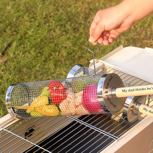 May include: A stainless steel grilling basket containing a variety of vegetables, such as red peppers, onions, and yellow courgettes. The basket has wooden handles with the text "Best chef & Best dad" and "My dad thinks he's".