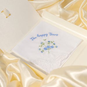 Könnte beinhalten: Weißes Taschentuch mit gestickten blauen Blumen und dem Text "For happy Tears". Das Taschentuch hat einen gewellten Spitzenrand und wird in einer cremefarbenen Schachtel auf einem Satin-Stoff präsentiert.