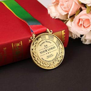 May include: A gold-colored medal with a red, green, and yellow ribbon. The medal has a laurel wreath design and text that reads "Event Name 1st YOUR LOGO -2025-". The medal is resting on a red book.