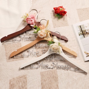 May include: Three wooden clothes hangers, each adorned with floral arrangements and personalized wire names. The hangers are in shades of brown, natural wood, and white. Each hanger has a different name and wedding date.