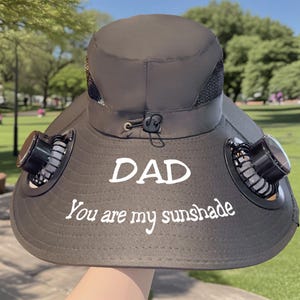 May include: A dark grey sun hat featuring two small fans on the brim. The hat has the text "DAD You are my sunshade" in white. Designed for sun protection and cooling, ideal for outdoor activities.