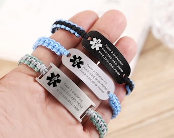 May include: Three medical alert bracelets with braided bands in black, blue, and green. Each bracelet features a rectangular metal plate with a medical symbol and text indicating Type 2 Diabetes and emergency contact information.