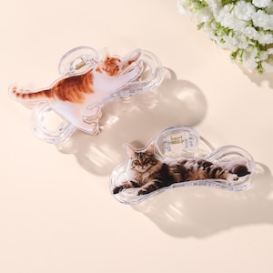 May include: Two clear plastic hair claw clips. One clip features a ginger and white cat stretching. The other clip has a brown and white cat lying down. Both clips have a spring mechanism.