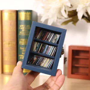 May include: A miniature blue bookshelf with three shelves filled with miniature books. The bookshelf is made of wood and has a rustic look.