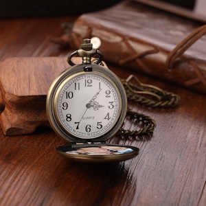 May include: Vintage bronze pocket watch with a white face, black numerals, and a quartz movement. The watch has a chain and opens to reveal a small image. The watch is on a wooden surface.
