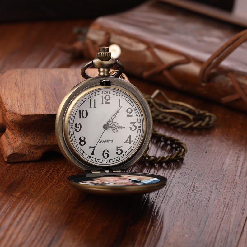 May include: Vintage bronze pocket watch with a white face, black numerals, and a quartz movement. The watch has a chain and opens to reveal a small image. The watch is on a wooden surface.