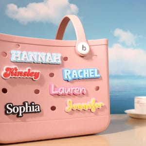 May include: A pale pink tote bag with name charms spelling out Hannah, Kinsley, Rachel, Lauren, Sophia, and Jennifer. The background shows a blue sky and sea. The bag has a pale pink handle.