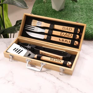 Peut inclure: Un ensemble d'ustensiles de barbecue en bois dans une boîte en bambou. L'ensemble comprend un pinceau, une fourchette, un couteau, une spatule et des pinces. Les manches sont gravés des mots "DAD", "MUM" et "KEVIN". La boîte a une poignée et des fermoirs argentés.