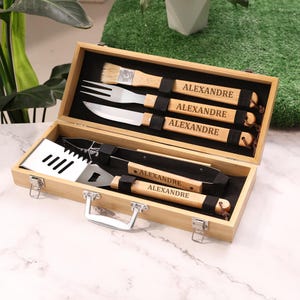Può includere: Un set di utensili da barbecue in bambù in una scatola di legno. Il set include un pennello, una forchetta, un coltello, una spatola e delle pinze. Ogni utensile ha un manico in legno con il nome "ALEXANDRE" inciso.