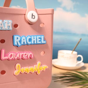 Könnte beinhalten: Eine hellrosa Tragetasche mit einem Griff mit dem Buchstaben "b". Die Tasche zeigt Namensanhänger: "Rachel" in Blau, "Lauren" in Rosa und "Jennifer" in Gelb. Eine weiße Teetasse und Untertasse sind im Hintergrund zu sehen.