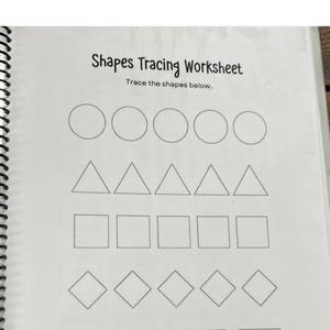 May include: A spiral-bound worksheet with the title "Shapes Tracing Worksheet." The worksheet features rows of circles, triangles, squares, and diamonds for tracing. The background is a wooden surface.