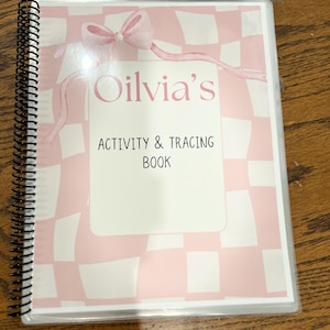 May include: A spiral-bound activity and tracing book with a pink and white checkered cover. The cover features the name "Oilvia's" in pink script, a pink bow, and the text "ACTIVITY & TRACING BOOK" in black.