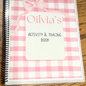 May include: A spiral-bound activity and tracing book with a pink gingham pattern cover. The cover features a white rectangle with the name "Olivia's" and the text "Activity & Tracing Book" in black font, and a pink bow.