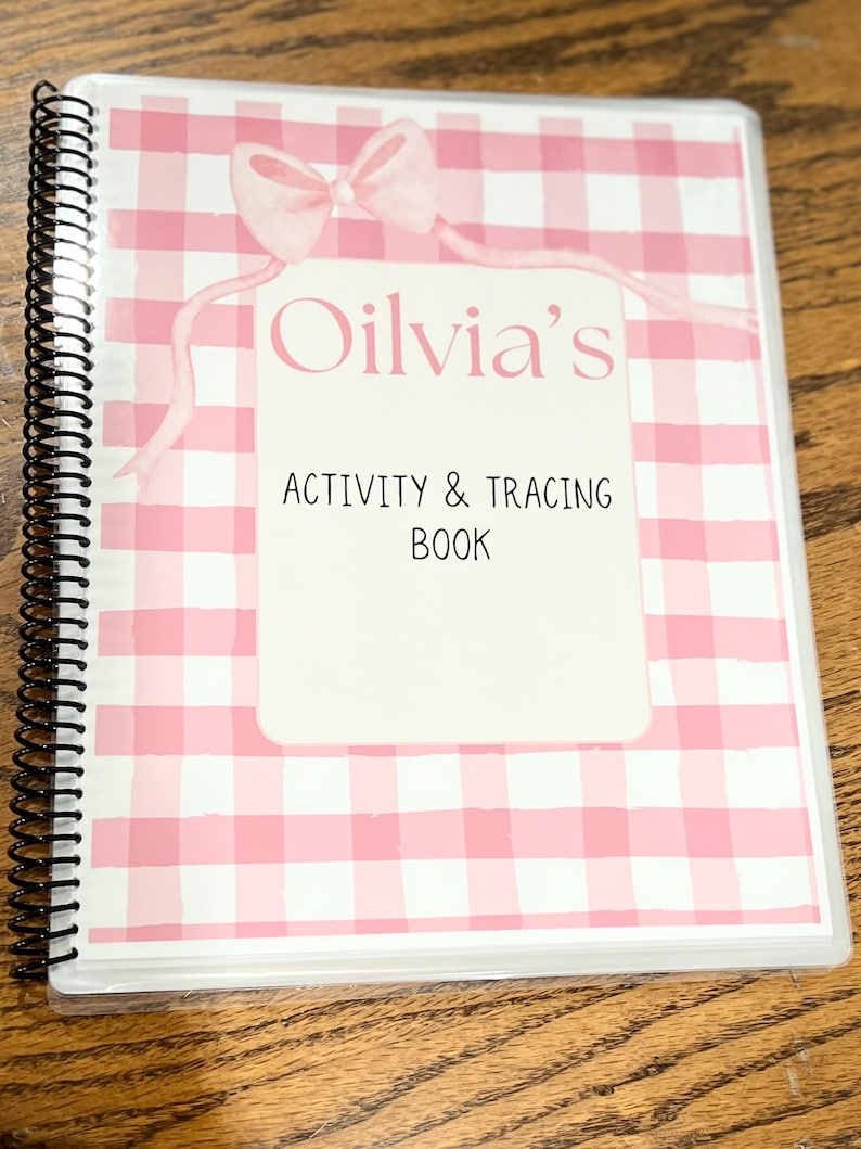 May include: A spiral-bound activity and tracing book with a pink gingham pattern cover. The cover features a white rectangle with the name "Olivia's" and the text "Activity & Tracing Book" in black font, and a pink bow.
