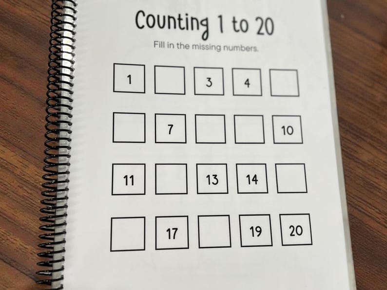 May include: A spiral-bound workbook page with the title "Counting 1 to 20." The page features a grid of squares with some numbers filled in, and blank spaces for children to complete the sequence. The workbook is on a wooden surface.
