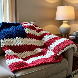 Puede incluir: Una manta gruesa tejida a mano, que recuerda a la bandera estadounidense, sobre un sofá. La manta presenta rayas rojas y blancas con un cuadrado azul en la esquina. Un elemento de decoración del hogar patriótico. El sofá está en un salón.