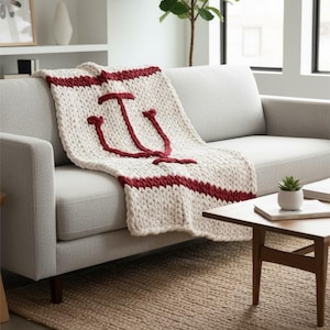 Op de afbeelding: Crèmekleurige, dik gebreide plaid met een grote bordeauxrode letter "I" en bordeauxrode strepen. De plaid is over een grijze bank gedrapeerd. De plaid is ongeveer 152 cm bij 127 cm.