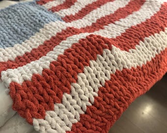 May include: A cozy, hand-knitted blanket featuring a design reminiscent of the American flag. The blanket showcases alternating stripes of red and white, with a blue square in the corner. The chunky knit adds texture and warmth to this patriotic home decor item.