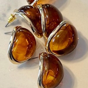 May include: Close-up of several amber-colored, teardrop-shaped earrings with gold-colored metal accents. The earrings have a post-back closure. The earrings are on a white surface.