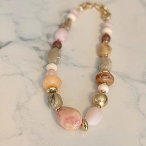 May include: A necklace featuring a variety of beads in shades of pink, peach, brown, and gold. The beads are strung together with a gold-colored chain. The necklace has a mix of round and irregular shaped beads.