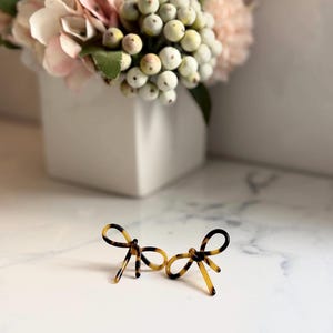 May include: A pair of bow-shaped earrings with a tortoiseshell pattern. The earrings are made of a translucent material with brown and black swirls. The bows are displayed on a white surface, with a vase of flowers in the background.