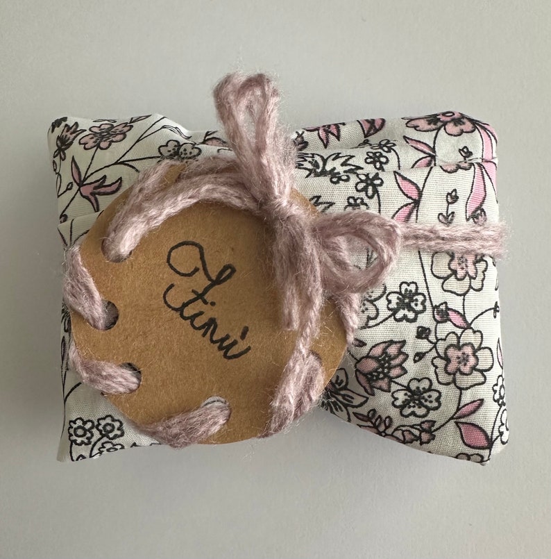May include: A small, white fabric pouch with a pink floral pattern. The pouch is tied with a pink ribbon and has a brown tag with the word "Tini" written on it.