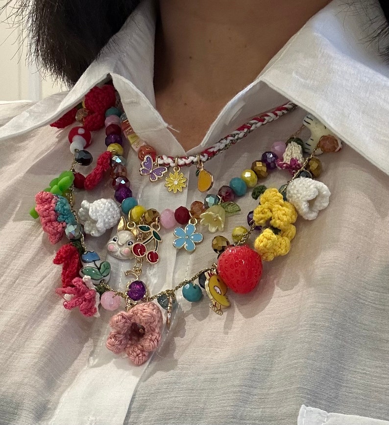 May include: A multi-layered necklace with a variety of colorful charms, including crocheted flowers, a bunny, a cherry, a strawberry, and a butterfly. The necklace is made of beads, string, and metal.