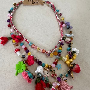 May include: Multi-strand necklace with a variety of colorful beads, charms, and crocheted flowers. The necklace features a small crocheted bunny, a mushroom, a four-leaf clover, and a strawberry charm. The tag on the necklace reads "Ten&uuml;."