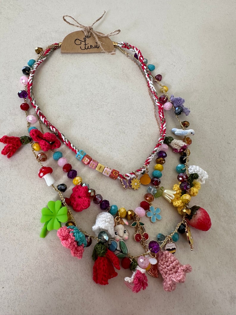 May include: Multi-strand necklace with a variety of colorful beads, charms, and crocheted flowers. The necklace features a small crocheted bunny, a mushroom, a four-leaf clover, and a strawberry charm. The tag on the necklace reads "Ten&uuml;."