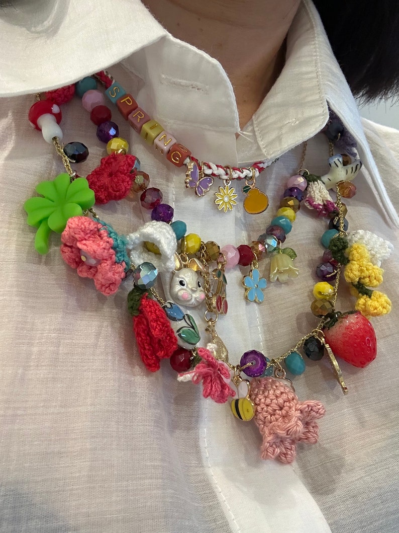 May include: A multi-layered necklace with a variety of charms, including crocheted flowers, a bunny, a strawberry, a bee, and a butterfly. The necklace also features a beaded section with the word "SPRING" spelled out in colorful beads.