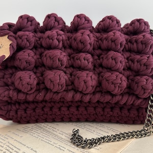 Op de afbeelding: Een bordeauxrode gebreide handtas met een kettingband. De tas heeft een textuur met bubbels. De tas ligt op een boek met het woord "Lezioni" zichtbaar op een label dat aan de tas is bevestigd.