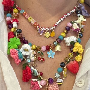 May include: A multi-layered necklace with a variety of charms, including crocheted flowers, a bunny, a strawberry, a cherry, and a bee. The necklace also features a beaded section with the word "SPRING" spelled out in colorful beads.