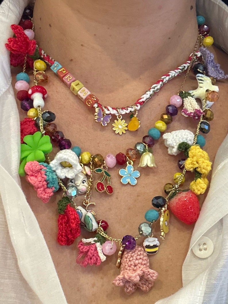 May include: A multi-layered necklace with a variety of charms, including crocheted flowers, a bunny, a strawberry, a cherry, and a bee. The necklace also features a beaded section with the word "SPRING" spelled out in colorful beads.